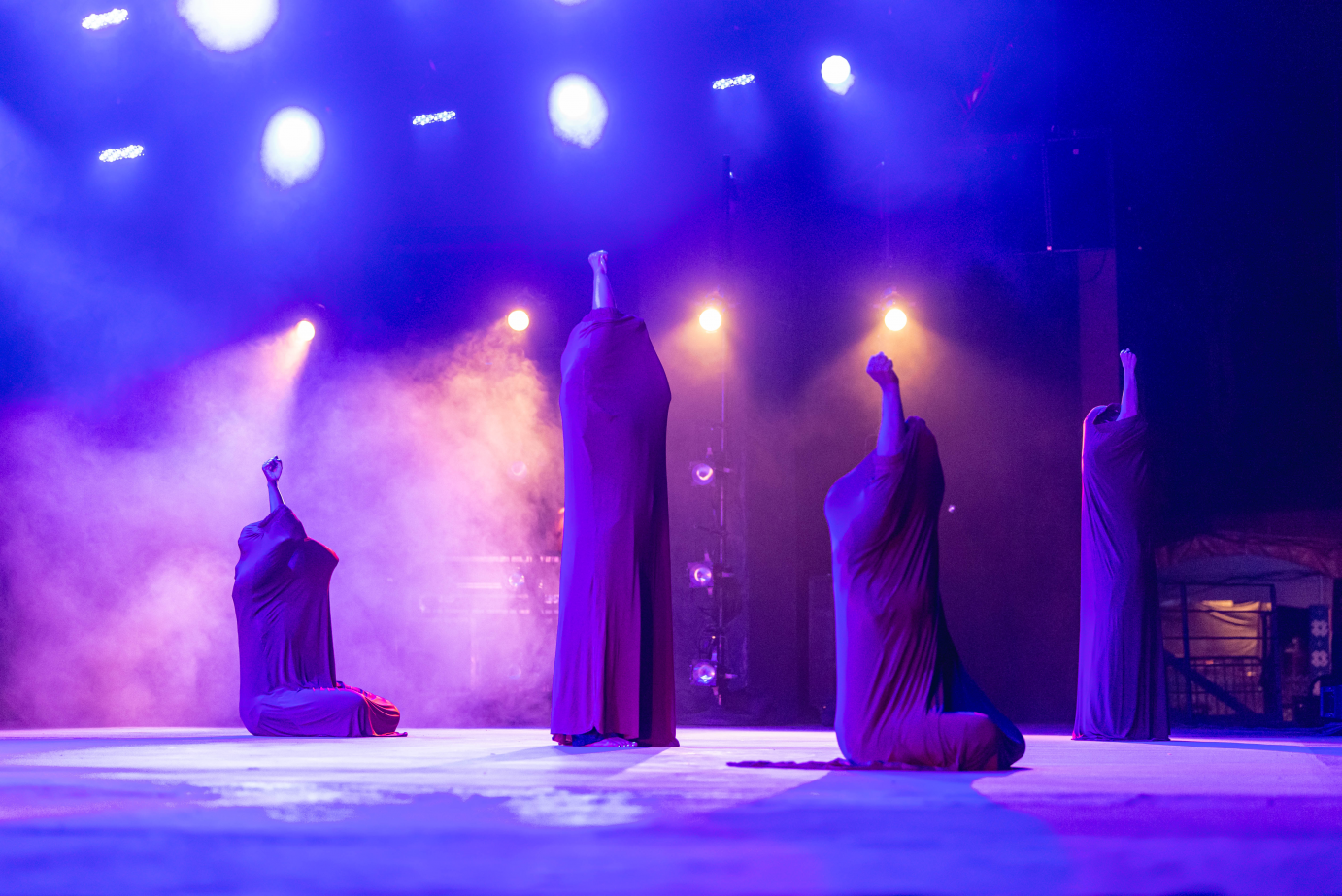 Four dancers are encased in tubes of purple fabric as they extend clasped hands to the sky. Lights stream from above them.